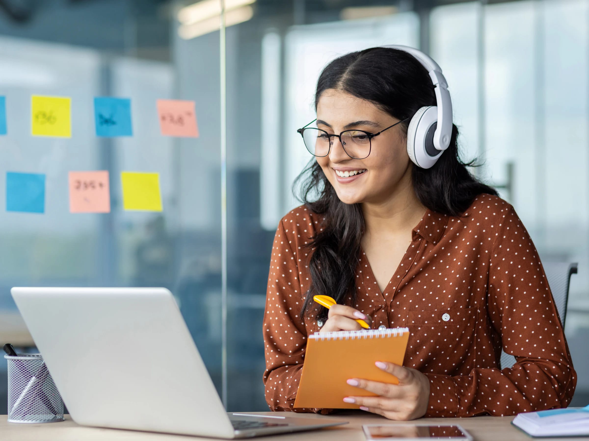 Mujer joven con lentes y audífonos blancos sonríe frente a su laptop mientras toma notas en un cuaderno durante una clase online